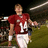 Pixelated image of a person in a red jersey with white number 11, white pants, and white socks standing on a grassy field with blurred background elements.