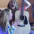 A person with long light brown hair plays a white acoustic guitar, wearing a blue and white patterned top, with a blurred background.