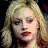 Pixelated static image of a person with light-colored hair styled in waves, wearing dark eye makeup and red lipstick against a black background.