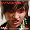 A person with dark hair and red eyeshadow wears a beaded necklace, mouth open in an expressive pose