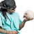 A person in a light blue shirt and dark bandana holds a white skull prop, with one hand resting on their stomach.