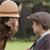 Two pixelated figures face each other outdoors; one wears a light hat with a brown band, the other a gray cap, both in period attire against a blurred green and dark background.