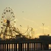 A static image of a Ferris wheel and roller coaster silhouetted against a sunset sky with a wooden pier railing in the foreground.