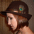 Pixelated image of a person wearing a dark brown wide-brimmed hat with a small green and blue feather, light skin tone, and long brown hair against a muted brown background.