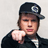 Static pixelated image of a person wearing a dark cap with a circular emblem, black jacket over white shirt, and making a thumbs-up gesture against light gray background.