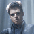 Pixelated image of a person with dark hair styled upward, wearing a dark jacket over a light shirt against a blue-gray background.