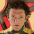 Pixelated close-up of a person with dark curly hair, wearing a dark jacket with green accents and a light-colored shirt, set against a yellow and red background.