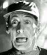 Black-and-white static image of a man's face with exaggerated expression, wearing a cap and high-collared shirt against blurred background.