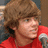 Pixelated image of a person with light brown curly hair wearing a red shirt, holding a silver microphone against a blurred background.