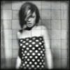 Black-and-white static image of a person with short dark hair wearing a strapless dress with white square patterns on a black background, standing against a grid-patterned backdrop.