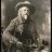 Pixelated grayscale image of a person wearing a wide-brimmed hat and dark clothing, seated with arms crossed against a muted background.