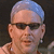 Static pixelated image of a person with short light-colored hair, dark sunglasses, and visible facial features against a dark background.