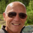 Pixelated close-up of a bald person wearing dark sunglasses and a black-collared shirt against green foliage background.