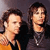 Two people with brown hair stand side-by-side against a dark background, one wearing a yellow top and the other a dark top with a silver necklace.