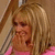 A woman with shoulder-length blonde hair in a pink top covers her mouth while smiling, set against a light brown background.