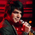A person with dark hair holds a silver microphone against a red striped background, wearing a dark jacket over a red shirt.