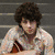 A static image of a person with curly brown hair in a light-colored shirt holding an acoustic guitar against gray steps.