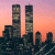 Pixelated static image of two tall skyscrapers against a gradient sunset sky with orange and pink hues, surrounded by smaller buildings in the foreground.