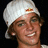 Pixelated close-up of a person wearing a white cap with small red and orange emblem, smiling against dark background.