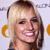 Pixelated close-up of a smiling person with blonde hair, blue eyes, and pink lips, wearing a dark top with visible orange circular shapes on the sides.