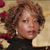 Pixelated portrait of a person with curly brown hair wearing a black top and silver earrings against a beige background with red floral elements.