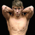 A shirtless man with short brown hair stands against a black background, arms raised behind his head in a pose.