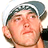 Pixelated close-up of a person wearing a dark cap with white trim, holding a microphone near their mouth against a plain background.