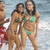 Three women in bikinis stand on a beach with ocean waves behind them; one woman wears red shorts.