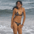 A woman in a black bikini stands in shallow ocean water, facing forward with a slight smile, her right hand resting on her hip.