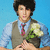 A person with dark hair wears a light blue shirt, gray vest, and red tie while holding white flowers against a blue background.