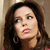Pixelated close-up of a person with long dark brown hair, light skin, and a neutral expression, wearing a dark top with a visible red shoulder strap.