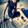 A person in a dark sleeveless dress with a light necklace sits on a sandy surface, hand touching water creating concentric ripples.