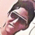 Pixelated close-up of a person's face with dark hair, wearing dark sunglasses, and a slight smile against a light background.