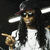 A person with long dark hair wears a dark cap with red trim, white shirt, and sunglasses, pointing right with a slight smile against a dark background with bright horizontal lights.