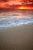 Pixelated sunset over water with orange and red sky, blue-gray waves, and brownish shore; small red dots on sand.