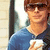A person in a blue shirt holding a white object with brown accents, wearing sunglasses and standing against a light-colored brick wall background.