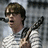 Pixelated image of a person with short brown hair wearing a white shirt, holding a black microphone near their mouth against a gray background.