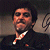Pixelated static image of a man in formal attire with a gold chain, set against a dark background with light gradient.