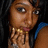 Pixelated close-up of a person with brown skin tone, dark eyes, and dark hair, wearing a blue garment and gold jewelry, hand near face.