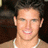 A pixelated image of a person with short dark hair wearing a light brown shirt and white necklace, smiling with visible teeth against a dark background.