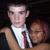Two people in formal attire pose closely together against a dark background, one wearing a white shirt and red tie.