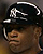Pixelated close-up of a person wearing a black cap with white curved lines and a stylized white logo on the front, set against a dark background.