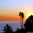 Pixelated sunset scene with orange and yellow sky, dark silhouettes of trees or structures on water's edge.