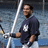 A person in a dark blue shirt with a white logo holds a bat, standing in front of a blurred background with vertical white poles.