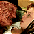 Two figures in profile view share a kiss, one with brown hair and the other with light brown hair, against a dark background with a light-colored object below.