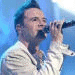 Four distinct stage performance images of a male singer in various outfits and poses holding a microphone against different colored backgrounds.