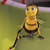 Static 50x50 pixel icon of a yellow cartoon bee with black stripes, blue eyes, and a smiling expression standing on green grass under a purple sky.