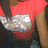 A person wearing a red short-sleeve shirt with silver graphic design, dark pants, and long brown hair; the lower right corner shows a gray area.