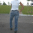 A person in a white shirt and blue jeans walks away from the camera on a paved path with green grass and trees in the background.