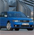 A static pixelated image of a blue sedan car with visible headlights and a front grille, positioned on a road with blurred background elements.